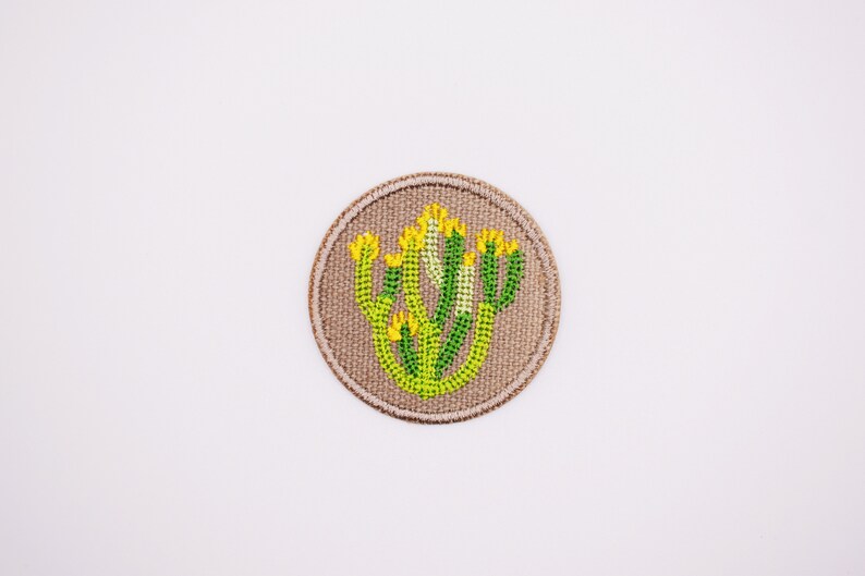 May include: A round, beige fabric patch with an embroidered cactus design in green and yellow.