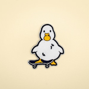 Puede incluir: Parche blanco bordado de un pato con un pico amarillo montando una patineta azul.