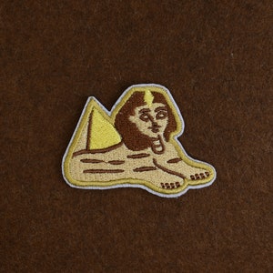 May include: Embroidered patch featuring the Sphinx of Giza. The patch has a tan body, brown hair, and a yellow headdress. The Sphinx is in a reclining position with its paws extended. The patch is outlined in white.