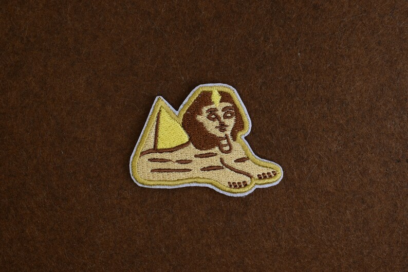 May include: Embroidered patch featuring the Sphinx of Giza. The patch has a tan body, brown hair, and a yellow headdress. The Sphinx is in a reclining position with its paws extended. The patch is outlined in white.