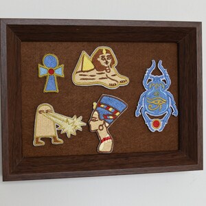May include: A framed display featuring embroidered patches with Egyptian motifs. The patches include an ankh, a sphinx, a scarab beetle, and a depiction of Nefertiti. The patches are in shades of blue, gold, and brown, set against a brown felt background.
