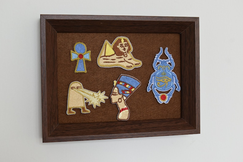 May include: A framed display featuring embroidered patches with Egyptian motifs. The patches include an ankh, a sphinx, a scarab beetle, and a depiction of Nefertiti. The patches are in shades of blue, gold, and brown, set against a brown felt background.