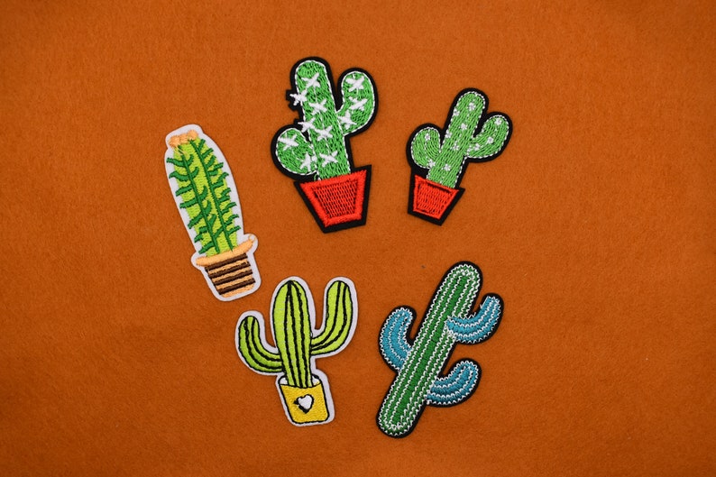 May include: Five embroidered cactus patches in different styles and colors. The patches are on a brown felt background.