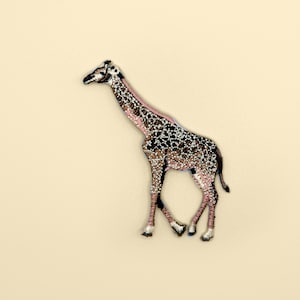 以下が含まれることがあります： 詳細なデザインのキリン型のブローチ。キリンは体に茶色と白の斑点模様、ピンク色の首、黒のアクセントがあります。ブローチはクリーム色の背景に配置されています。
