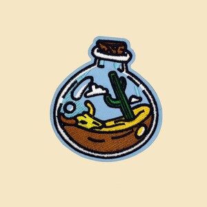 Könnte beinhalten: Eine blaue Glasflasche mit einem Korkverschluss, die eine Miniatur-Wüstenlandschaft mit einem Kaktus, Sand und Wolken enthält.