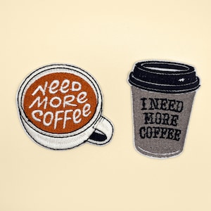 May include: Two embroidered iron-on patches featuring coffee mugs. One mug is white with brown coffee and the text "Need More Coffee". The other mug is grey with black text that reads "I Need More Coffee".