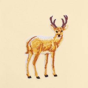 Könnte beinhalten: Ein braunes gesticktes Hirsch-Patch mit weißen Nähten und braunen Geweih. Der Hirsch steht auf einem hellgelben Hintergrund.