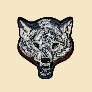 May include: Embroidered patch of a wolf's head with a snarling expression. The wolf has a gray and white fur pattern with brown accents around the muzzle and green eyes.