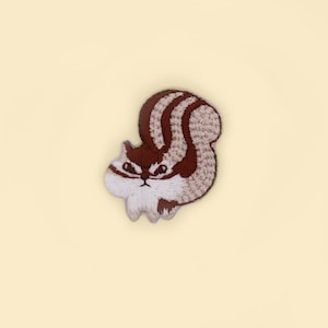 Peut inclure: Patch brodé représentant un écureuil brun et blanc avec une expression renfrognée.