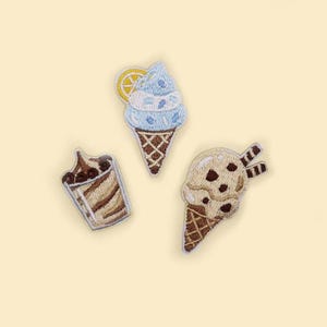Puede incluir: Tres parches bordados de helados. Uno es un cono de helado azul con una rodaja de limón, otro es un sundae de chocolate y el tercero es un cono de helado con chispas de chocolate con dos barquillos.