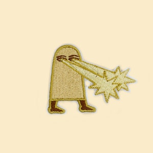 May include: Embroidered patch featuring a cartoon figure with a beige body and brown feet. The figure is shooting golden starbursts from its eyes. The patch has a beige background.