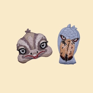 May include: Two embroidered patches featuring animal faces. One is a light brown, stylized lizard face with large eyes and a red mouth. The other is a blue and brown Shoebill bird head with an intense expression.