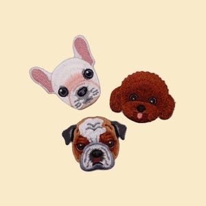 May include: Three embroidered dog face patches: a French Bulldog, a Poodle, and a Bulldog. The patches are in white, brown, and black, with detailed stitching. The background is a plain, light beige colour.