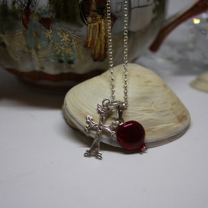 Puede incluir: Un collar de plata con un crucifijo y un dije de granada roja. El collar está sobre una superficie blanca con una concha y un jarrón de porcelana en el fondo.