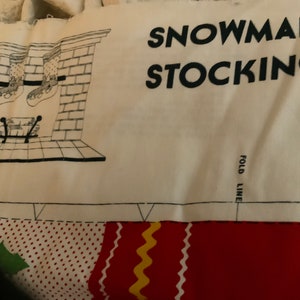 May include: A white fabric panel with black line drawings of a fireplace and stockings hanging from it. The text "SNOWMAN STOCKING" is printed in black at the top of the panel. The text "FOLD LINE" is printed in black at the bottom of the panel.