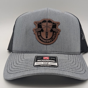 May include: A gray and black trucker hat with a brown leather patch featuring the 101st Airborne Division insignia. The patch has the words "De Oppresso Liber" in a circle around the insignia. The hat has a black mesh back and a black adjustable strap with the words "Richardson 112 Trucker" printed on it.
