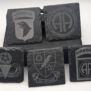 May include: Six black slate coasters with different military insignia designs. The coasters feature the 101st Airborne Division, 82nd Airborne Division, and the 173rd Airborne Brigade emblems. The coasters are engraved with the words "Airborne" and "Sua Sponte".