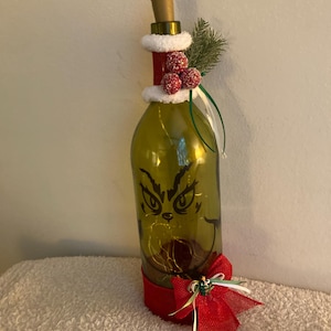 May include: A green glass bottle decorated with a black Grinch face decal, internal string lights, and a cork stopper. The neck features white faux fur trim and red berries, while the base has a red burlap ribbon with a small decorative bow.