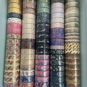 May include: A collection of washi tapes in a variety of colors and patterns, including gold, pink, purple, green, and black. The tapes are stacked in columns and feature designs such as stars, moons, hearts, and leaves.