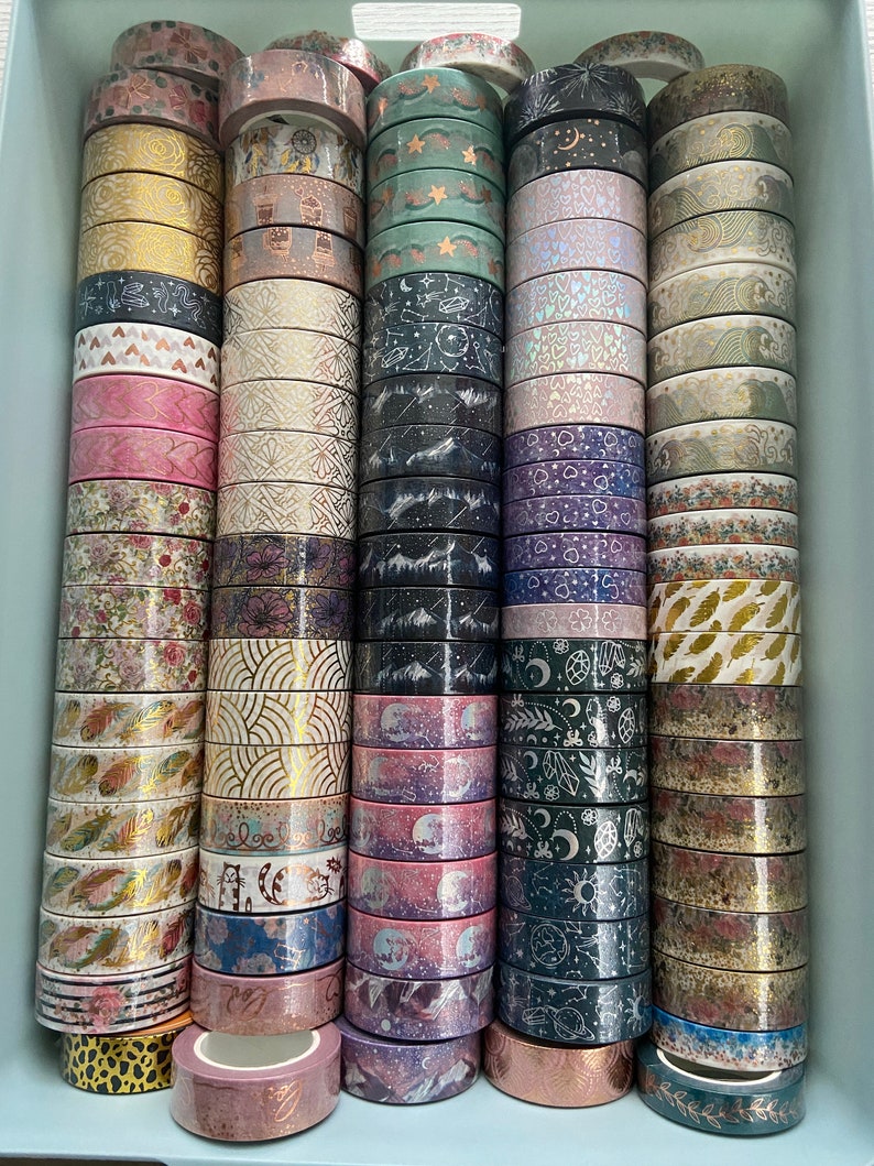 May include: A collection of washi tapes in a variety of colors and patterns, including gold, pink, purple, green, and black. The tapes are stacked in columns and feature designs such as stars, moons, hearts, and leaves.