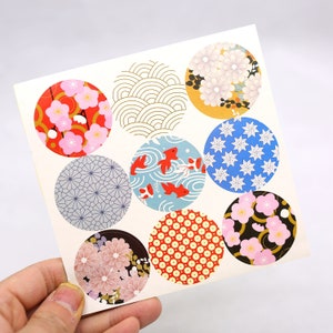 May include: A square sheet of paper featuring nine circular stickers with diverse patterns. Designs include red and pink plum blossoms, gold wave lines, white chrysanthemums on yellow, blue geometric stars, gray netting, red goldfish, and polka dots.