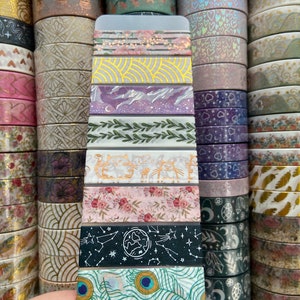May include: A close-up of a variety pack of washi tape featuring different patterns and colors, including floral, geometric, and abstract designs. The tape is displayed on a white card.