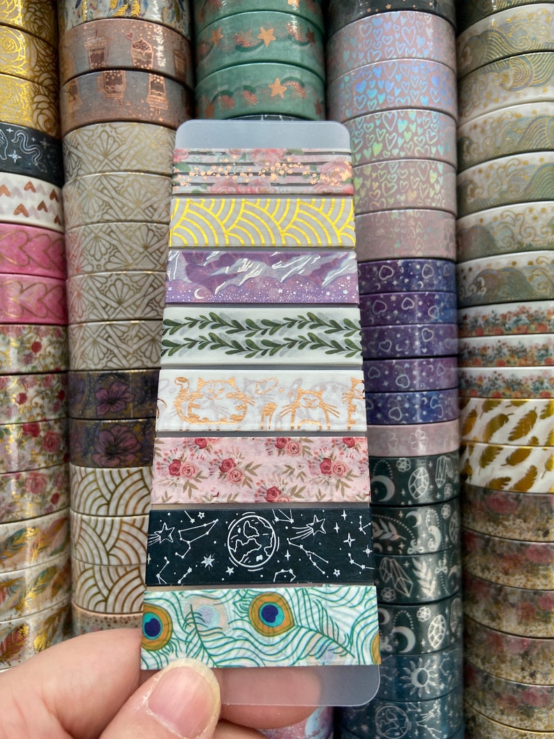 May include: A close-up of a variety pack of washi tape featuring different patterns and colors, including floral, geometric, and abstract designs. The tape is displayed on a white card.