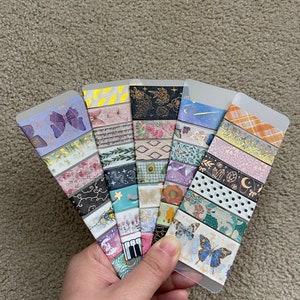 May include: A collection of 10 decorative washi tapes featuring various patterns, including butterflies, flowers, stars, and geometric designs. The tapes are arranged in a fan shape and are displayed on a light brown surface.