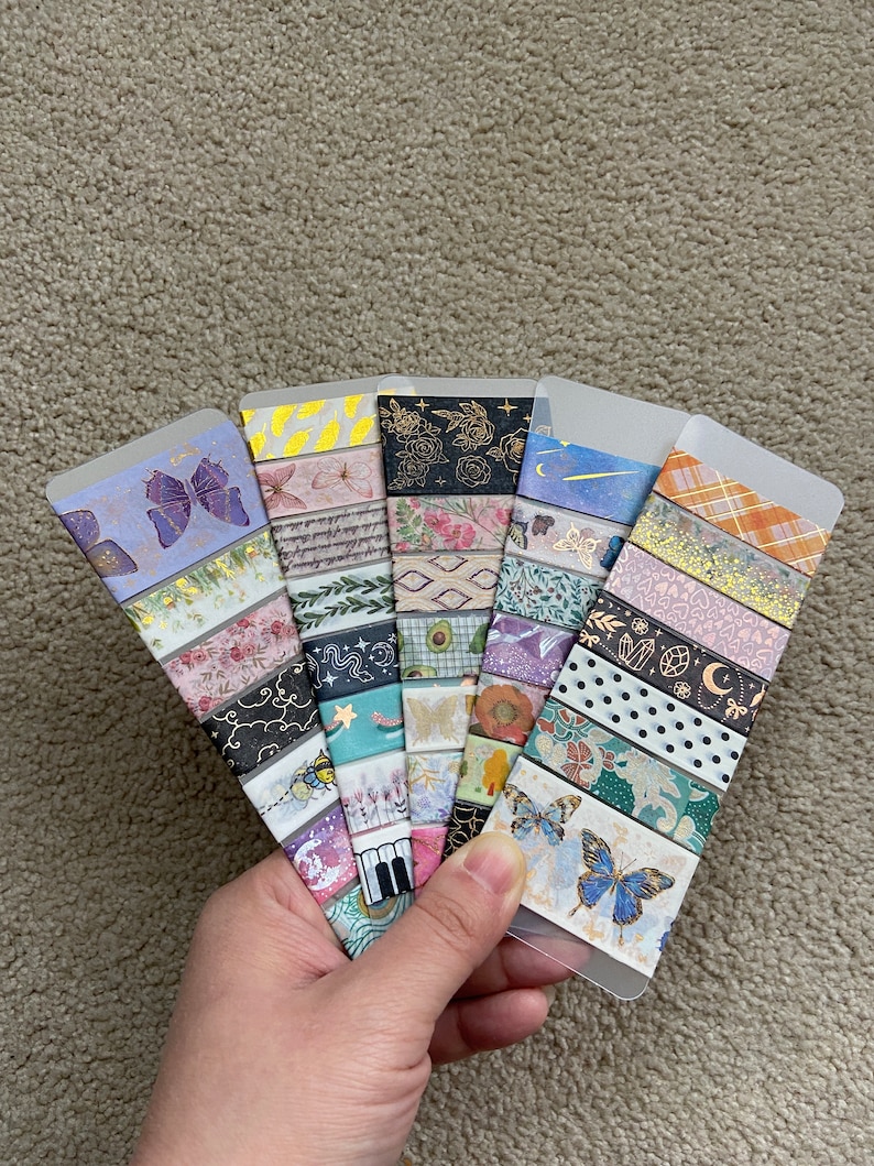 May include: A collection of 10 decorative washi tapes featuring various patterns, including butterflies, flowers, stars, and geometric designs. The tapes are arranged in a fan shape and are displayed on a light brown surface.