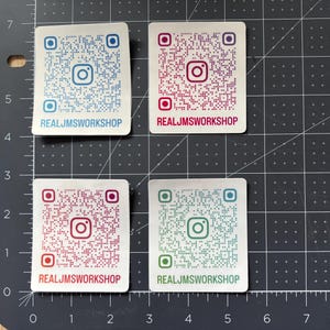 May include: Four rectangular stickers with rounded corners, each featuring a unique QR code and the Instagram logo. The text REALJMSWORKSHOP is printed at the bottom of each. The stickers are blue, magenta, red, and teal, set against a grid mat.
