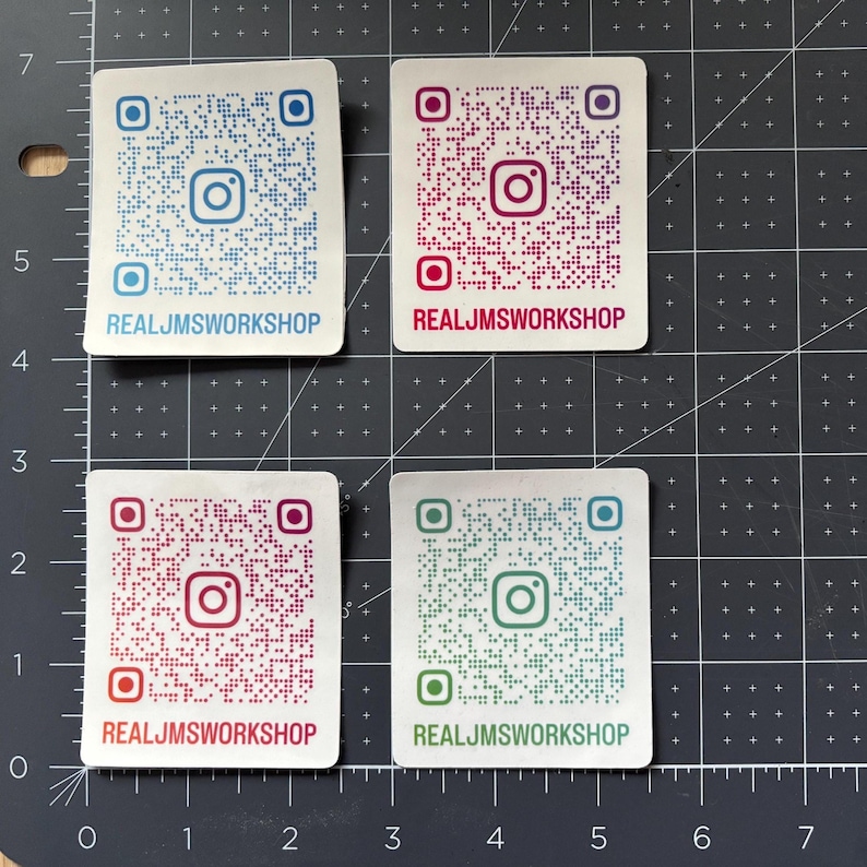 May include: Four rectangular stickers with rounded corners, each featuring a unique QR code and the Instagram logo. The text REALJMSWORKSHOP is printed at the bottom of each. The stickers are blue, magenta, red, and teal, set against a grid mat.