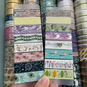 Peut inclure: Gros plan sur une variété de rubans washi, un matériau de bricolage populaire, avec différents motifs et couleurs. Les rubans sont disposés en grille et sont affichés sur une feuille de plastique transparente. Les rubans présentent des motifs tels que des fleurs, des étoiles, des lunes et des motifs géométriques.