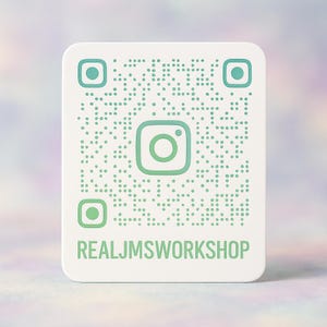 May include: A white square sign with rounded corners featuring a light green Instagram QR code and the text REALJMSWORKSHOP printed in light green capital letters at the bottom. The design is centered on a flat surface with a soft, light-colored background.