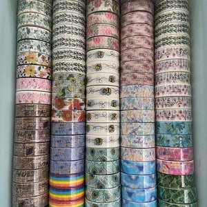 May include: A collection of colorful washi tapes with various patterns, including floral, geometric, and abstract designs. The tapes are stacked in a light blue container.