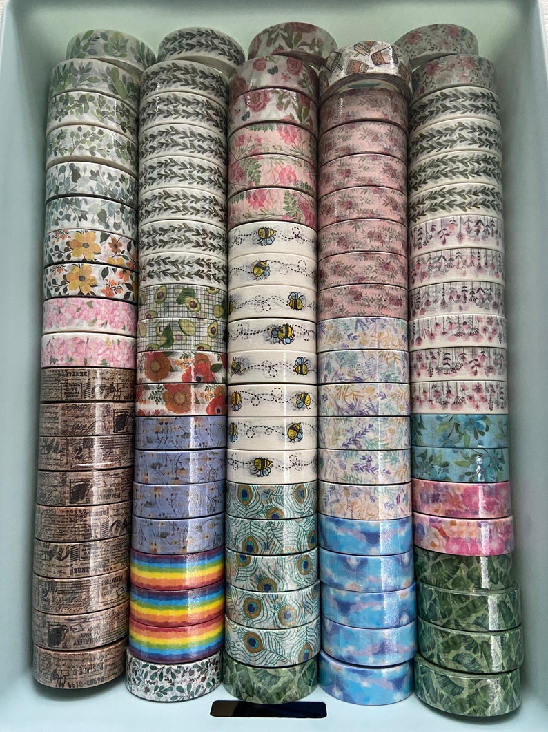 May include: A collection of colorful washi tapes with various patterns, including floral, geometric, and abstract designs. The tapes are stacked in a light blue container.