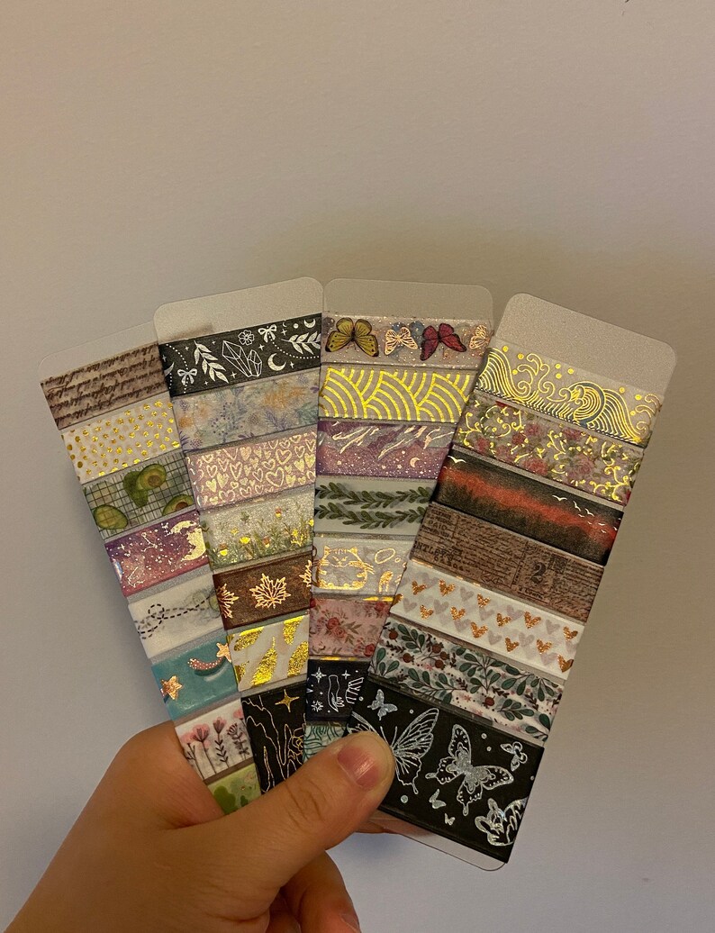 May include: A set of 10 decorative washi tapes featuring various patterns and designs, including butterflies, flowers, geometric shapes, and gold accents. The tapes are displayed on a clear plastic backing.