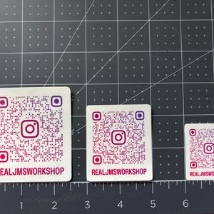 May include: Three square white stickers featuring a purple and pink Instagram QR code and the text REALJMSWORKSHOP in red. The stickers are arranged by size on a cutting mat with inch markings, showing widths of approximately 2.5, 2, and 1.5 inches.
