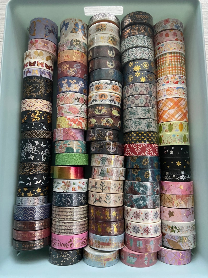 May include: A collection of colorful washi tapes in a light blue plastic container. The tapes feature various patterns, including floral, geometric, and animal prints.