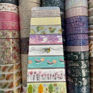May include: A close-up of a variety pack of washi tape featuring different patterns and colors, including avocado, stars, flowers, and a bee. The tape is displayed on a clear plastic card.