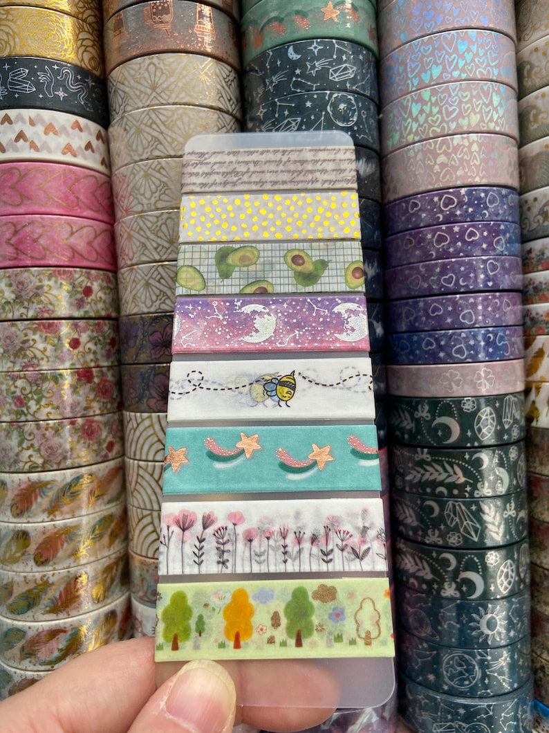 May include: A close-up of a variety pack of washi tape featuring different patterns and colors, including avocado, stars, flowers, and a bee. The tape is displayed on a clear plastic card.