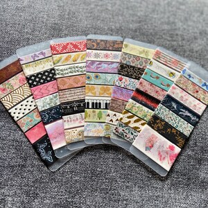May include: A collection of 15 decorative washi tapes with various patterns and colors, including floral, geometric, and abstract designs. The tapes are displayed on a gray background.