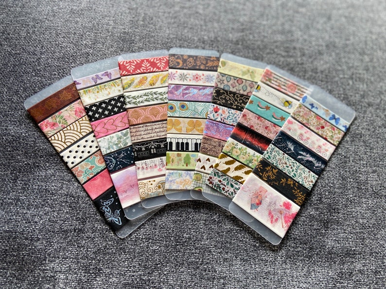 May include: A collection of 15 decorative washi tapes with various patterns and colors, including floral, geometric, and abstract designs. The tapes are displayed on a gray background.