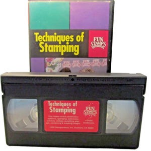 以下が含まれることがあります： 「Techniques of Stamping」というタイトルの黒いVHSテープ。赤いラベルにはFun Stampsのロゴとテキストが記載されています。背後には、緑と紫のブロックが描かれたカラフルなカバーが付いた、同じタイトルとロゴのVHSケースが立っています。