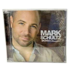 May include: A clear plastic CD jewel case featuring the album Broken & Beautiful by Mark Schultz. The cover art displays a portrait of a man with short hair and light stubble, wearing a light-colored collared shirt against a dark brown background.