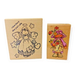 May include: Two rectangular wooden rubber stamps. The left stamp features a black line drawing of an angel with stars and the text Where love is there God is also. The right stamp features a colorful illustration of a person in a hat and dress.