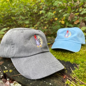 Peut inclure: Deux casquettes de baseball avec des patchs brodés d'une fusée qui décolle de la lune. L'une est grise et l'autre est bleu clair.