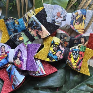 Puede incluir: Una colección de lazos para el cabello coloridos y brillantes con personajes de cómics como Superman, Wonder Woman y Wolverine. Los lazos tienen varios colores, incluyendo rojo, dorado, negro y morado, con detalles de purpurina.