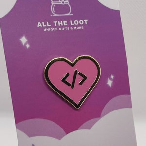 May include: Pink heart-shaped enamel pin with gold outline and code symbols inside. The pin is displayed on a small wooden easel with a purple background and stars. The background also has the text "All the Loot Unique Gifts & More".