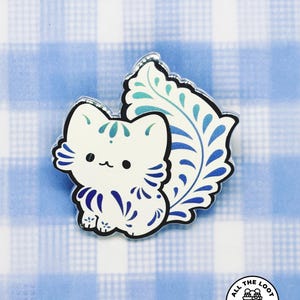 May include: A white cat-shaped pin with blue accents and a decorative blue and teal leaf design. The pin has a black outline and is set against a blue and white checkered background. The pin is a unique gift.