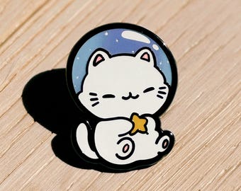 May include: A white cat with a yellow star in its paw sits inside a blue and white space-themed bubble. The pin is black and has a white outline. The pin is from the shop "All The Loot: Unique Gifts & More."